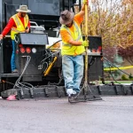 Large pavement truck dispersing black tar with two men smoothing the layed material