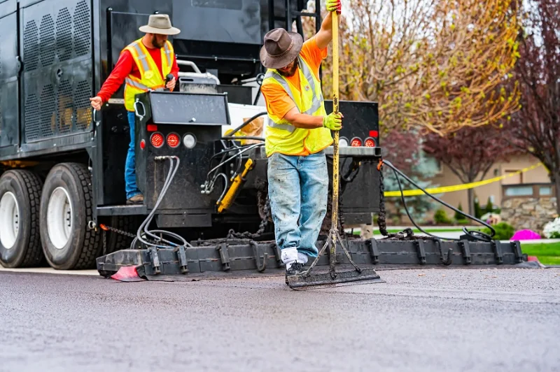 Large pavement truck dispersing black tar with two men smoothing the laid material
